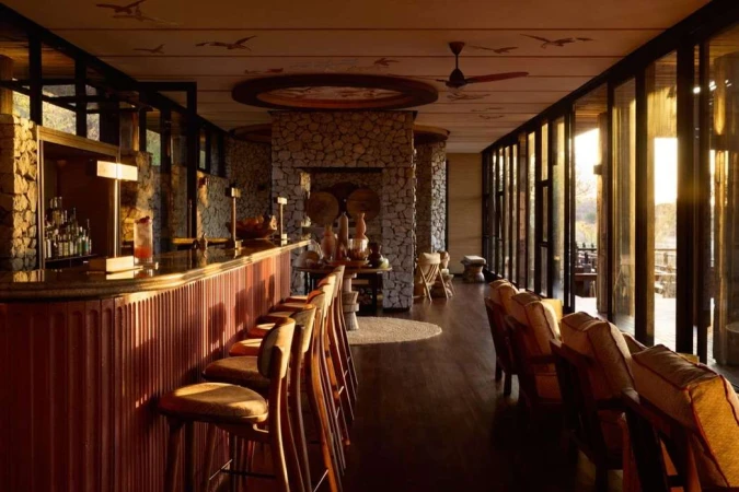 Interior of a hotel bar lounge area with stone walls, a bar with high chairs, tables with decorative items, and comfortable seating.