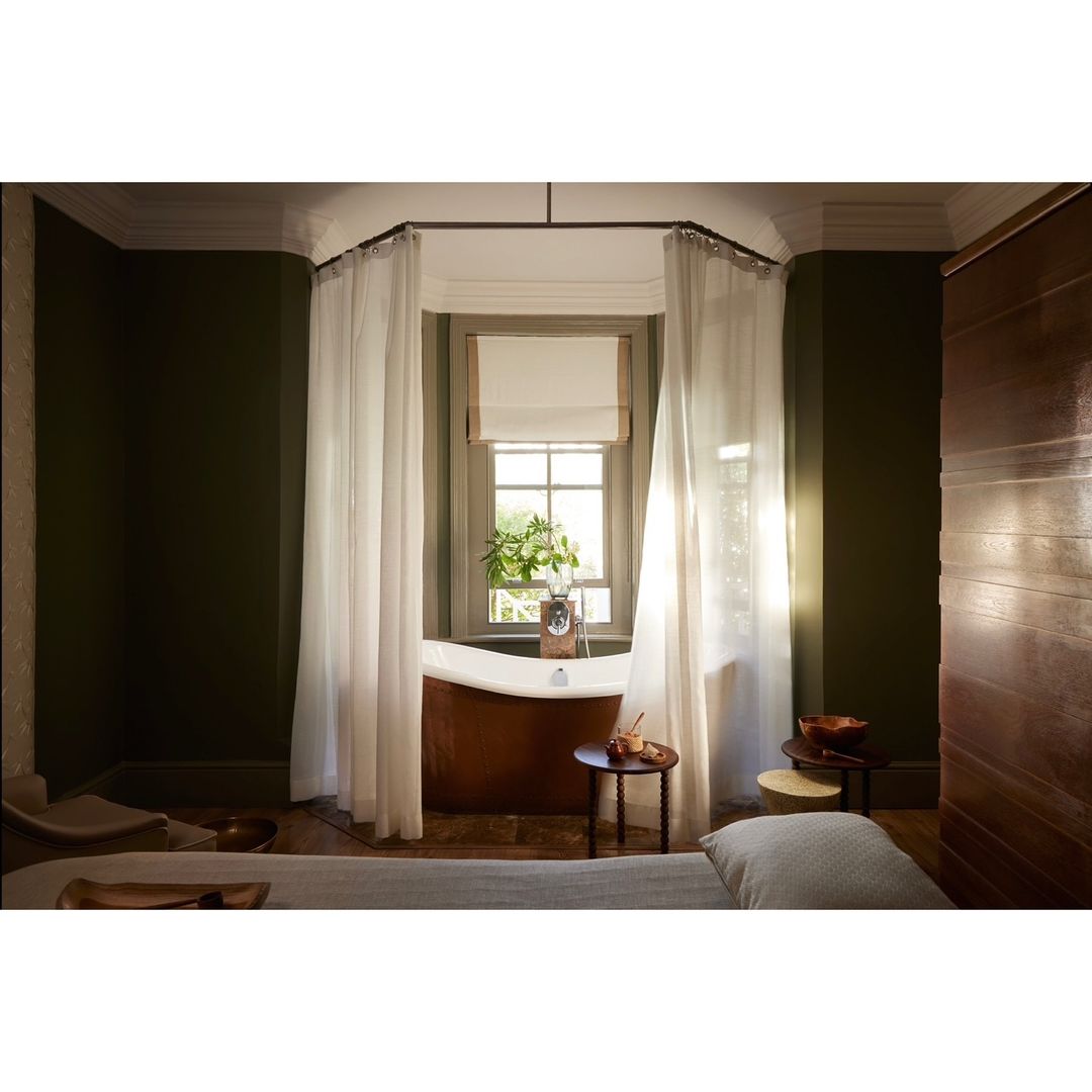 Interior view of a spa treatment room with a copper tub, round wood tables, sheer curtains, and dark green walls.