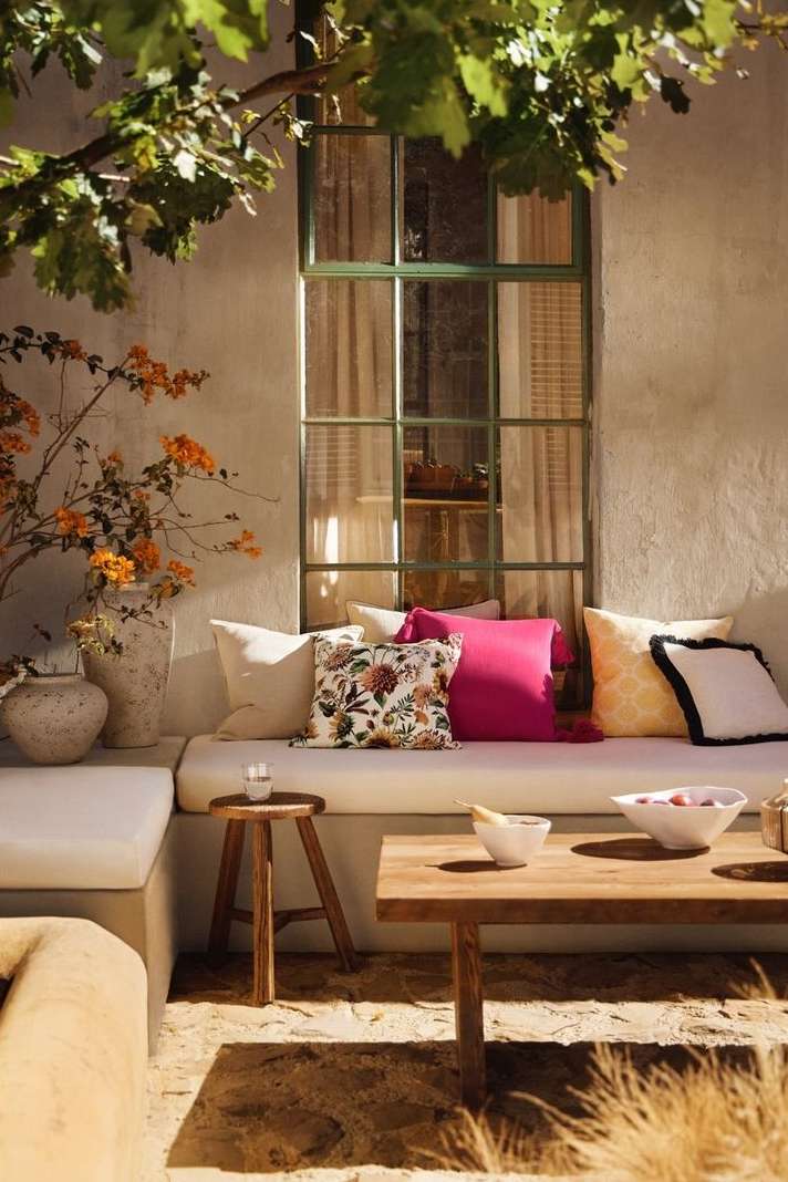 Sunlit outdoor seating area with neutral cushions, vibrant pink pillow, a wooden table and vase of orange flowers, creating a cozy, inviting space.
