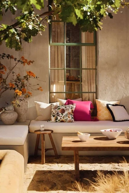 Sunlit outdoor seating area with neutral cushions, vibrant pink pillow, a wooden table and vase of orange flowers, creating a cozy, inviting space.