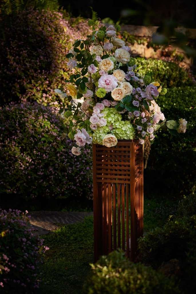 Floral arrangement with roses, hydrangeas, and other flowers in a brown wooden lattice vase in an outdoor garden setting.