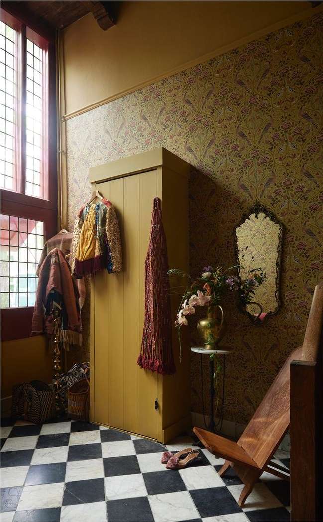 Eclectic hallway with vintage wallpaper, mustard-colored cabinet, tiled floor, ornate mirror, wooden chair, and hanging clothing.