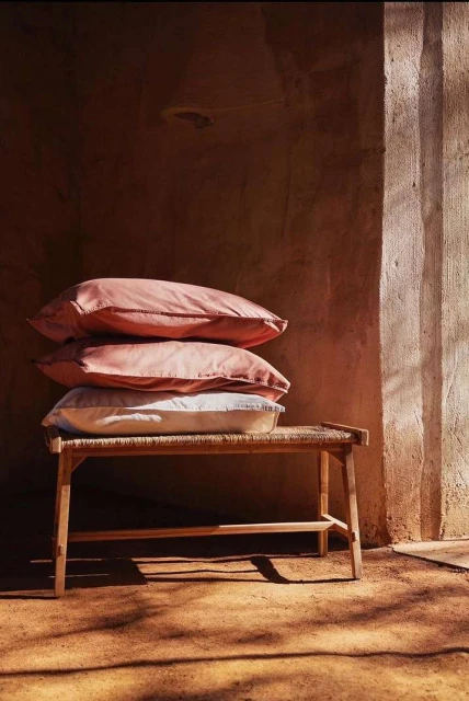 Stack of pillows on a wooden bench with woven seat, indoors against a textured wall with sunlight, creating a cozy interior setting.