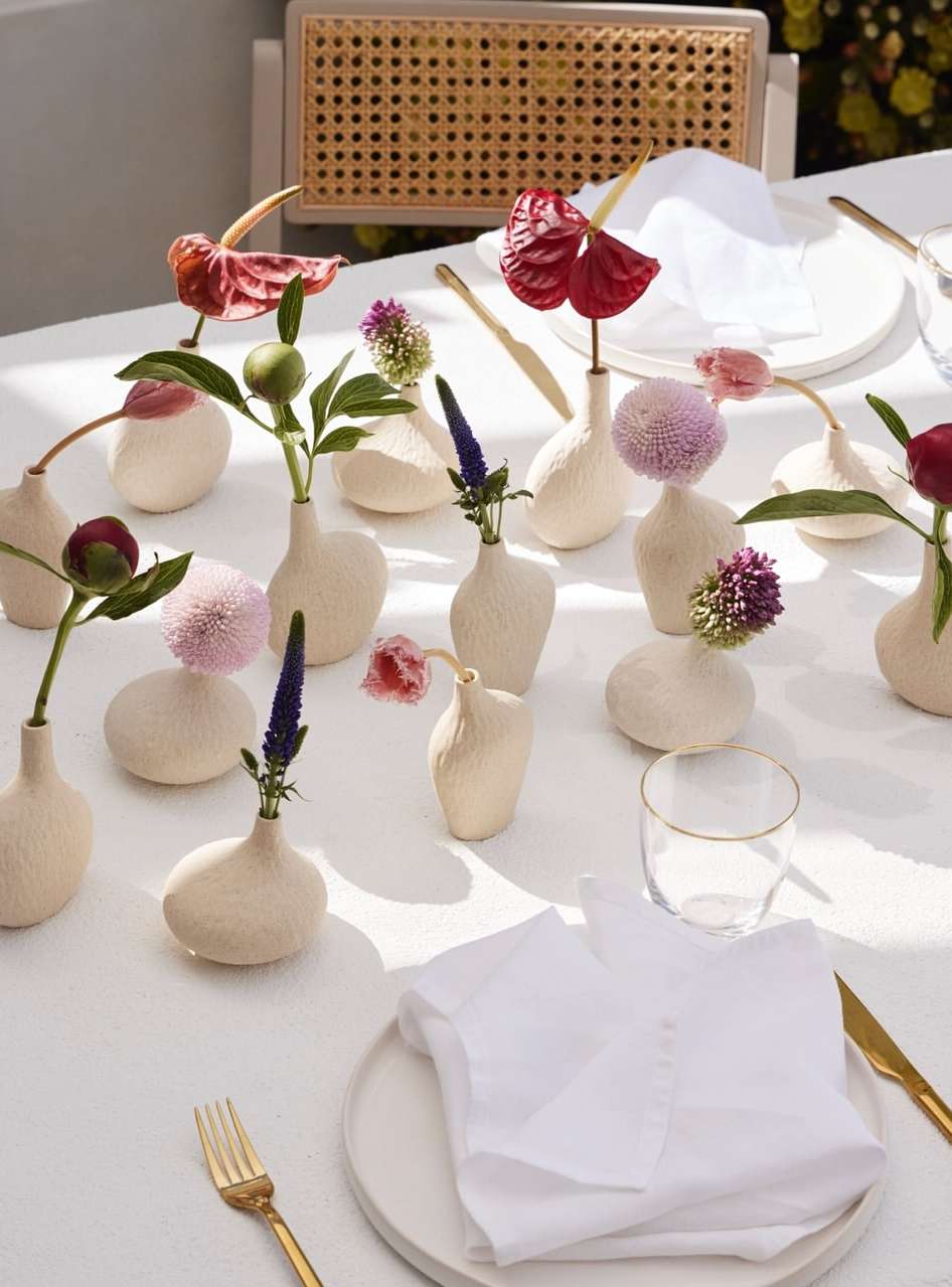 Table setting with white tablecloth, plates, gold flatware, and assorted flower arrangements in bud vases, creating an elegant floral tablescape.