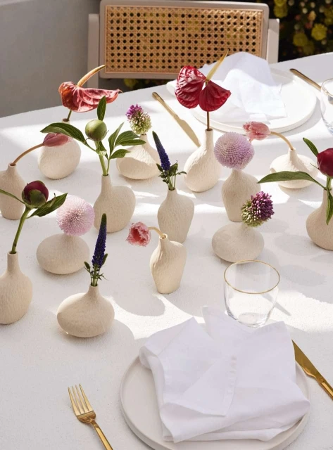 Table setting with white tablecloth, plates, gold flatware, and assorted flower arrangements in bud vases, creating an elegant floral tablescape.