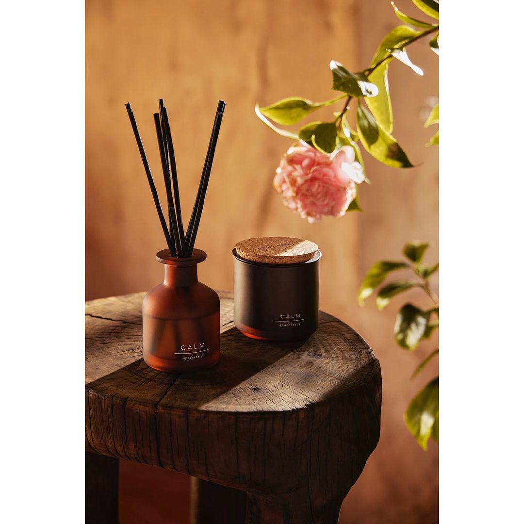 Aromatic reed diffuser and candle set with pink flowers on a wooden table, creating a calming and inviting ambiance in a warm-toned setting.