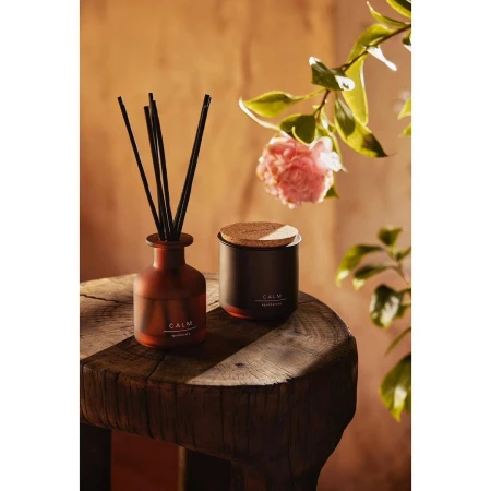 Aromatic reed diffuser and candle set with pink flowers on a wooden table, creating a calming and inviting ambiance in a warm-toned setting.