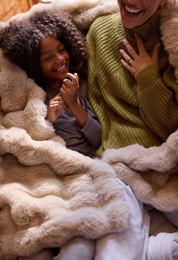 A smiling child with curly hair and a woman are covered in a fluffy blanket, indoors. They appear joyful and cozy together.