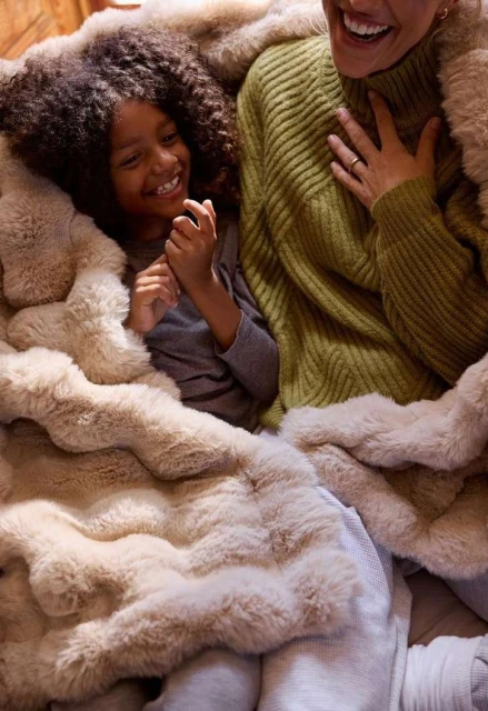 A smiling child with curly hair and a woman are covered in a fluffy blanket, indoors. They appear joyful and cozy together.