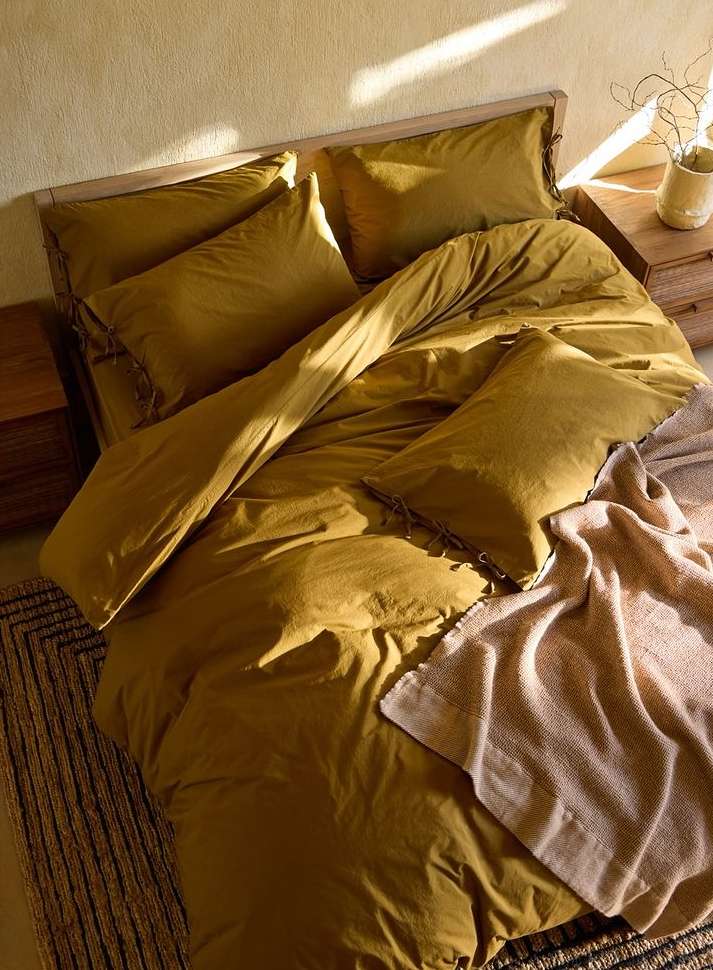 High-angle shot of a gold duvet cover on a bed with pillows, accompanied by a beige throw blanket and wooden nightstand.
