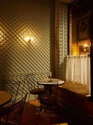 Interior cafe seating with wooden tables, metal chairs, lattice patterned wall panels, and a warm wall sconce lighting.