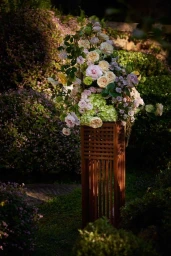 A lush arrangement of pastel garden roses and hydrangeas atop a tall wooden slatted stand outdoors in a garden setting.