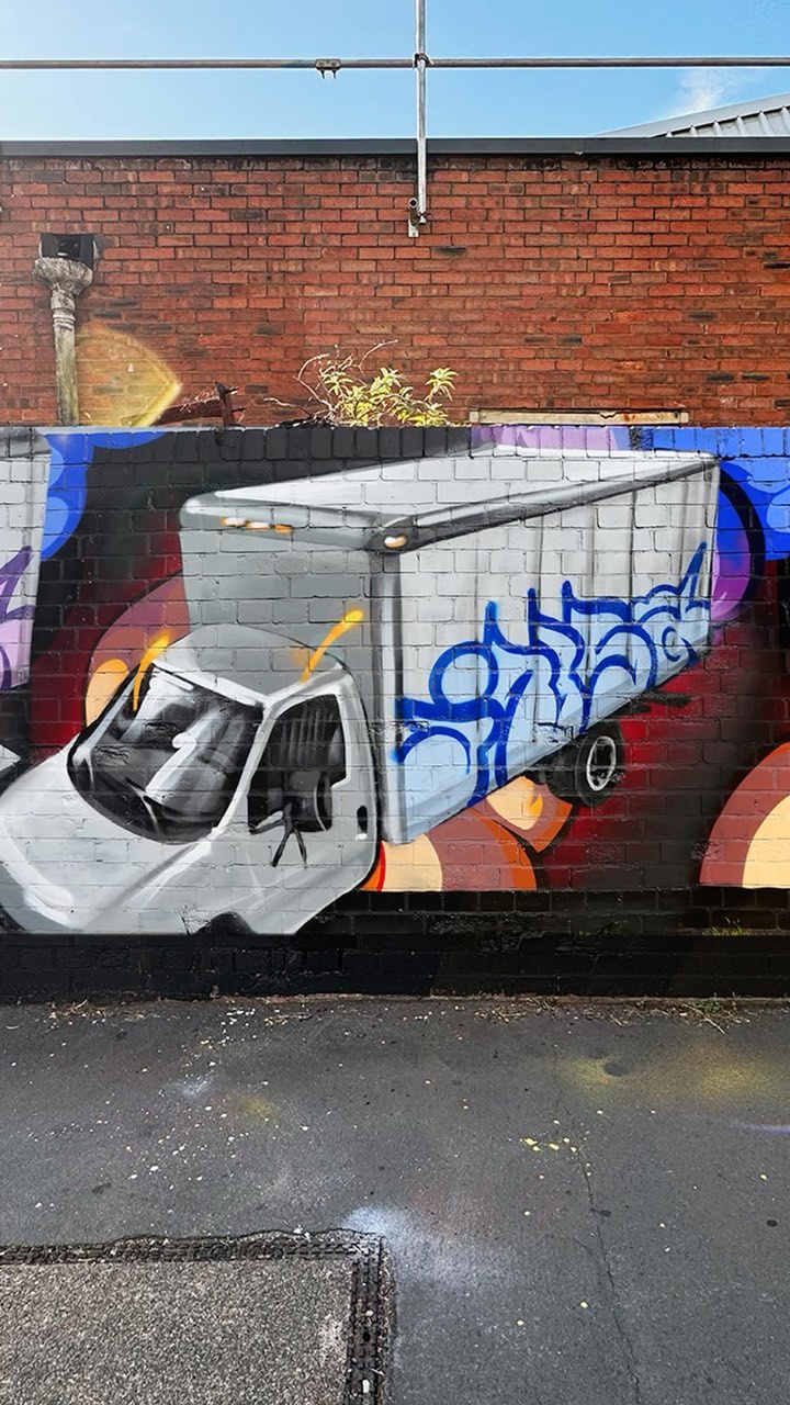 Graffiti art on a brick wall features a semi-truck with detailed shading and blue accents, complemented by abstract backgrounds.