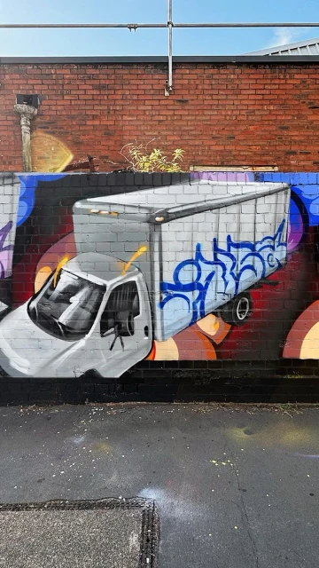 Graffiti art on a brick wall features a semi-truck with detailed shading and blue accents, complemented by abstract backgrounds.