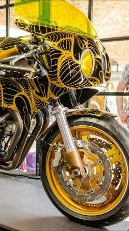 A close-up of a black and yellow custom motorcycle with intricate line details and a gold rimmed tire fills the frame.