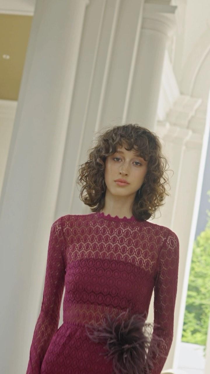 A woman with curly brown hair is posing in a purple lace dress with a feather accent in an outdoor setting featuring white columns.
