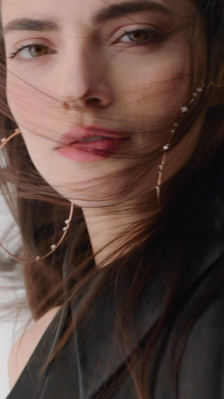 Close-up portrait of a young brunette woman with medium skin tone and light eyes, wearing large hoop earrings and a black top.