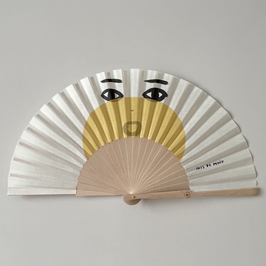 A hand fan with a playful face design dominates the image, set against a neutral backdrop, showcasing a blend of artistry and utility.