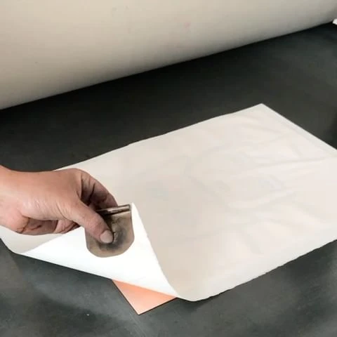 Hand lifting corner of white paper on a dark surface, revealing a pink surface underneath, with a metal tool on top of the paper.