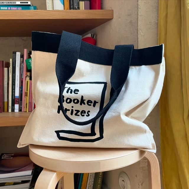 Canvas tote bag with black handles and 'The Booker Prizes' printed on the front sitting on a light-colored wooden stool.