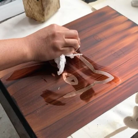 Close-up of a person's hand cleaning a wooden board with a cloth, revealing a stencil-like design on the dark wood surface.