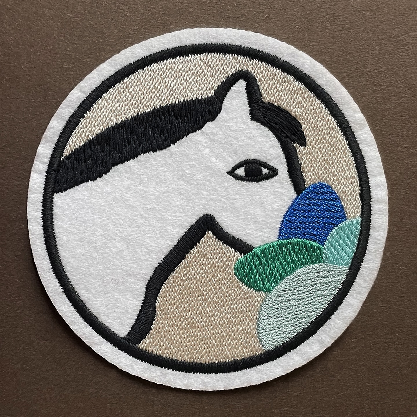 Embroidered patch of a stylized white horse head with black accents, set against a beige backdrop within a black and white circle.