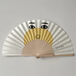 An open hand fan featuring a painted face illustration with dark eyes, eyebrows, and a yellow circular element by Iris De Mouy.