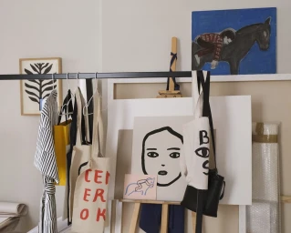 Illustrated tote bags and striped garment hanging on a black rack inside an artist studio with paintings in background