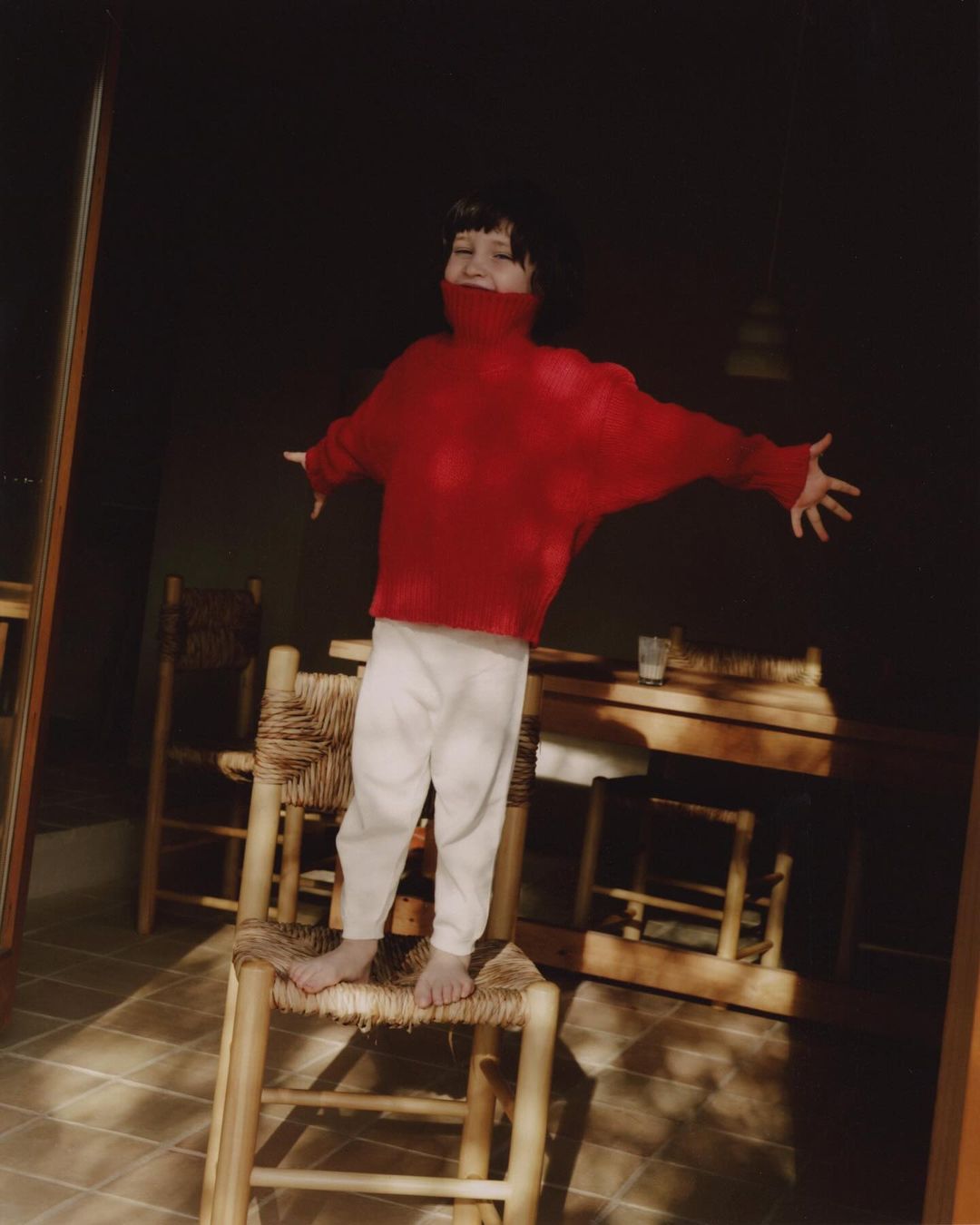 Child in red sweater stands on woven chair with arms out wide in sunlit room; table and chairs visible in background.