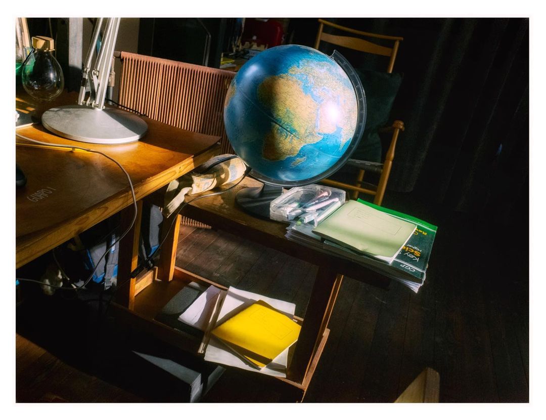 A world globe sits on a wooden table in a room with indirect sunlight. Additional items include a desk lamp and books, with a cozy atmosphere.