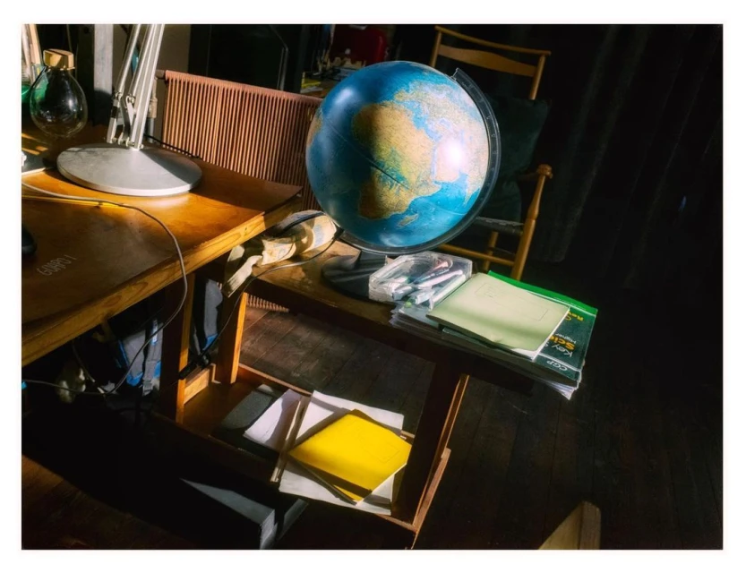 A world globe sits on a wooden table in a room with indirect sunlight. Additional items include a desk lamp and books, with a cozy atmosphere.