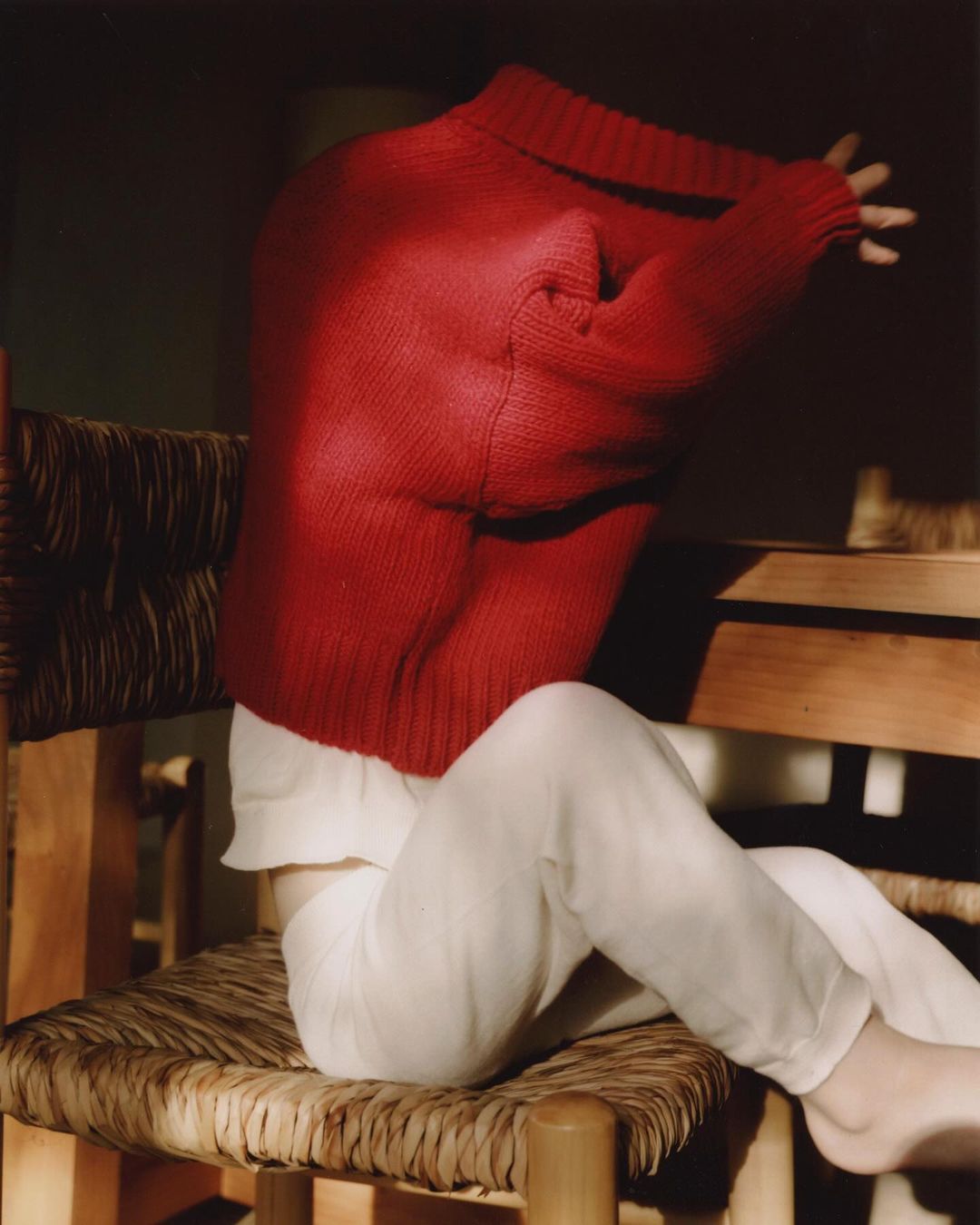Person in a red sweater sits on a woven chair, illuminated by sunlight, creating a cozy and intimate indoor atmosphere. 