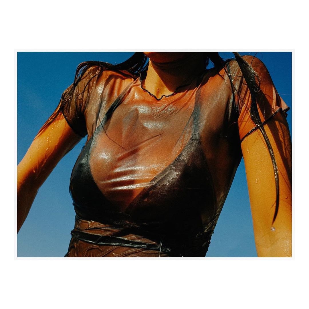 Young woman in wet, transparent mesh top and black bikini top against a bright blue sky, creating a fashion or beauty-focused image.