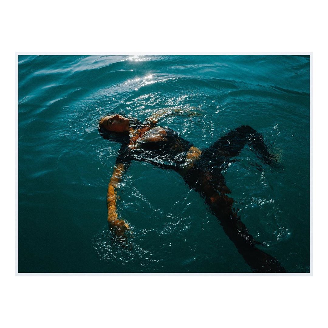Woman in dark swimsuit and leggings floats on her back in blue water with sunlight reflecting off the surface on a sunny day.