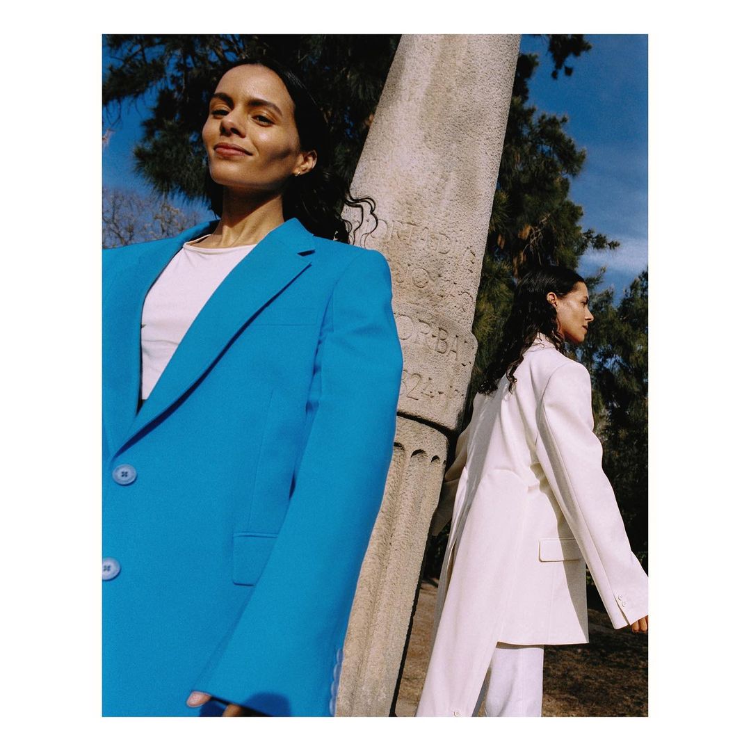 Two adult females pose in blazers, one blue and one white, next to a stone column outside on a sunny day. Fashion editorial.