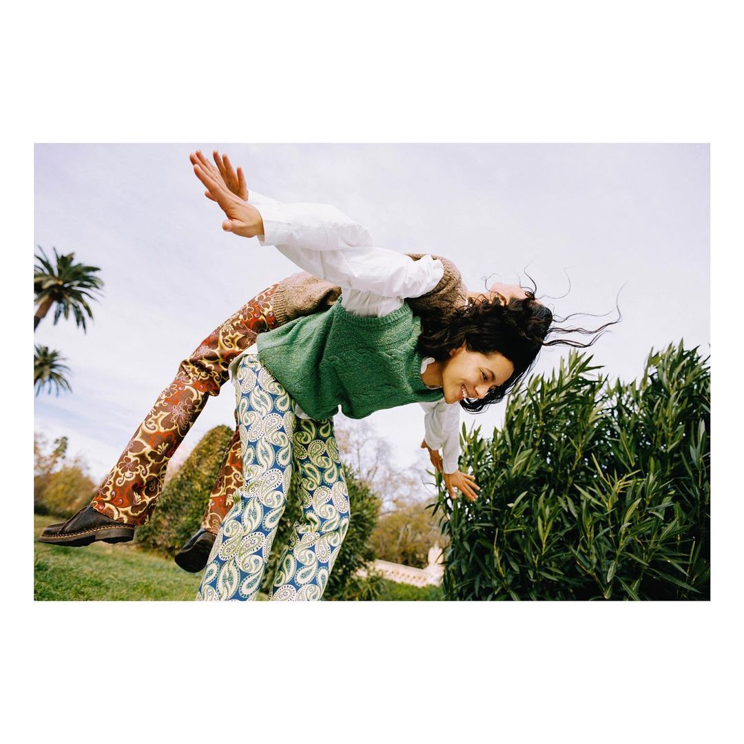 Two women outdoors in patterned pants, green top, and white shirt, playfully positioned against a bright sky, showcasing a carefree moment.