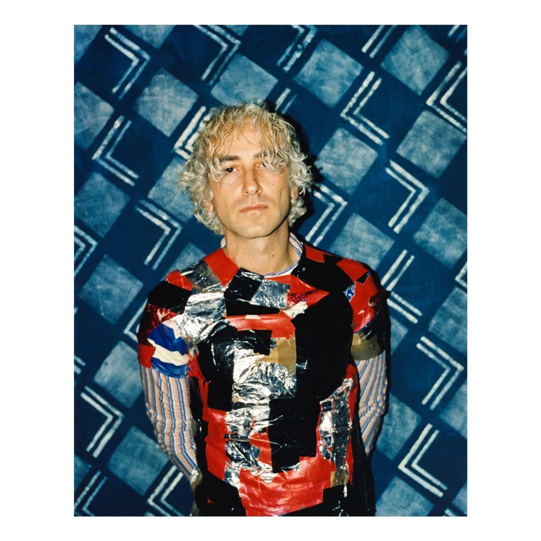 Portrait of adult male with wavy blonde hair wearing a brightly colored shirt with blue patterned background in an indoor setting.