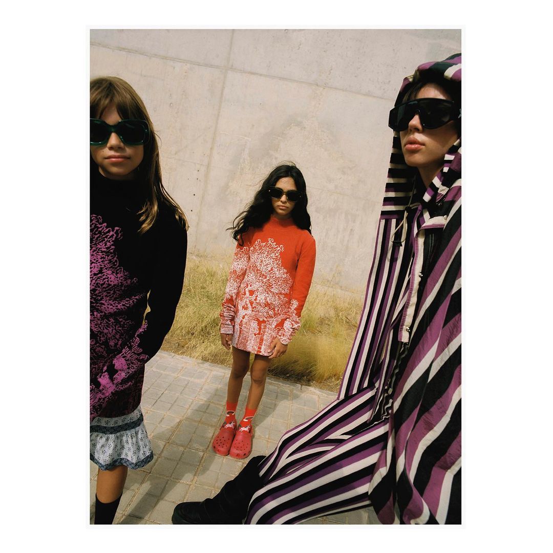 Three young people in a contemporary fashion shot wearing colorful outfits and sunglasses posing against a concrete wall outside during the day.