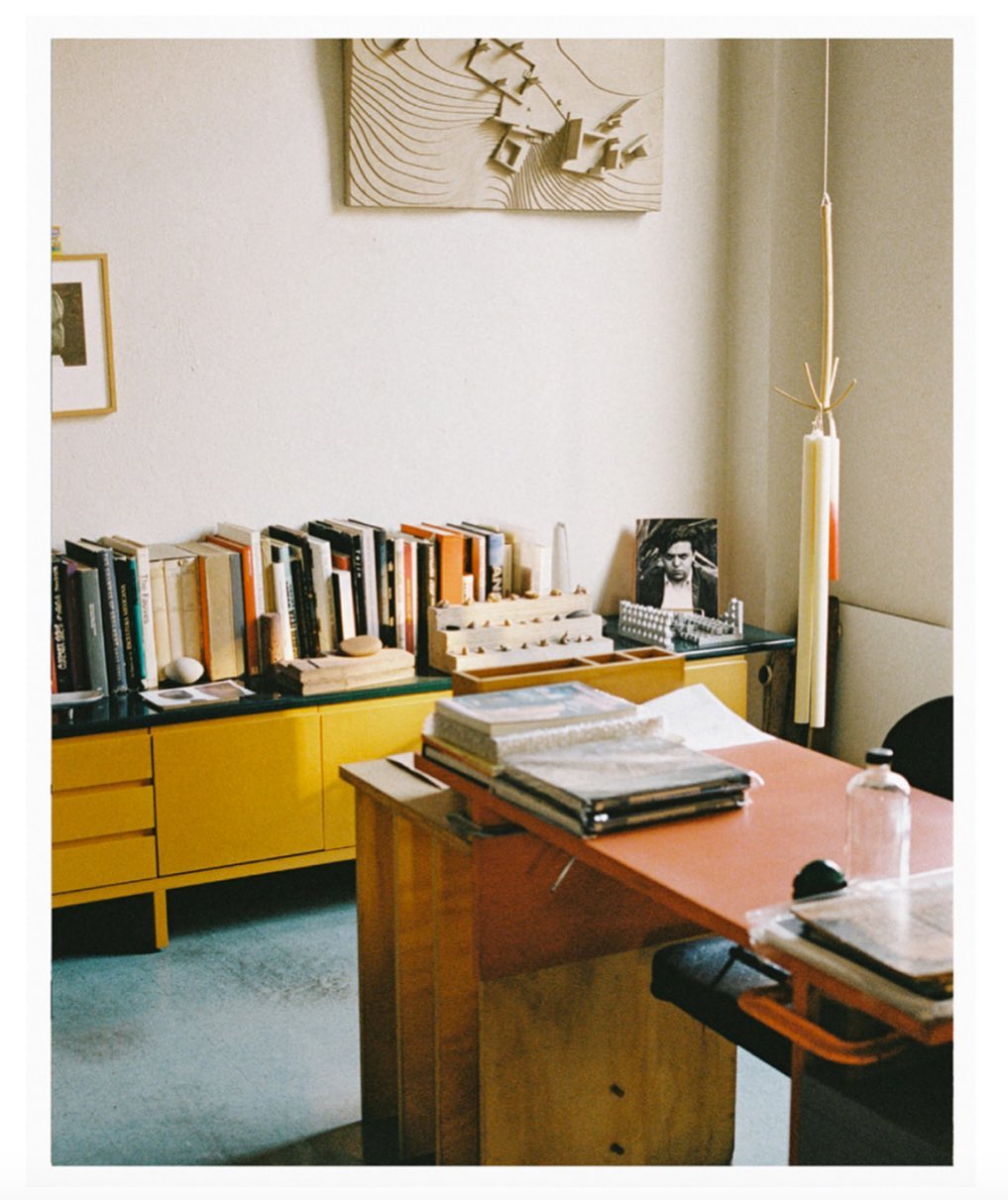 Interior view of a vintage office featuring a desk with books, a yellow cabinet, and architectural models in a softly lit room.