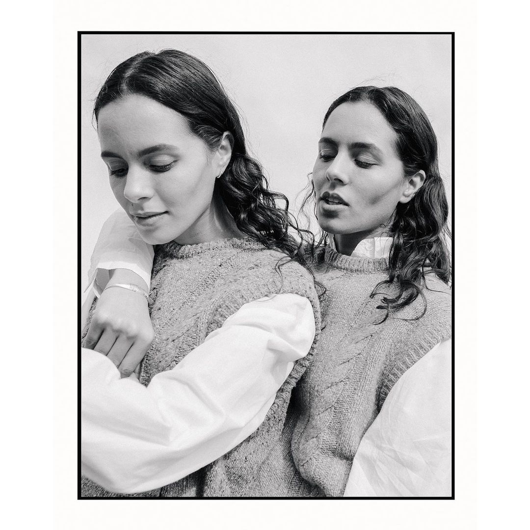 Monochromatic portrait of two women with curly brown hair wearing matching sweaters and white shirts in a close-up studio shot.