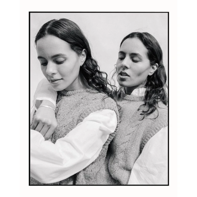 Monochromatic portrait of two women with curly brown hair wearing matching sweaters and white shirts in a close-up studio shot.