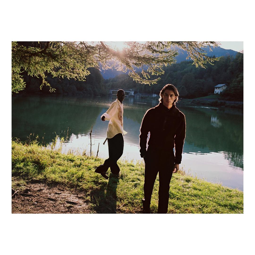 Two people stand on a grassy shore by a lake with mountains in the background, bathed in sunlight filtering through trees.
