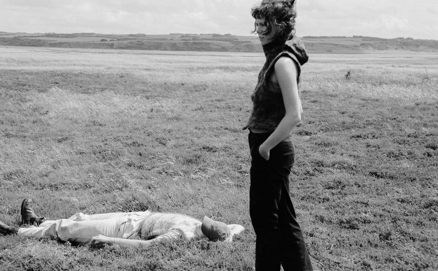Monochrome image of a woman standing near a man lying in a grassy field, under an overcast sky, with a distant treeline.