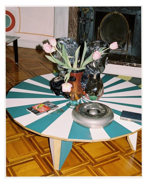 Still life composition: pink tulips in glass vases on a geometric-patterned round table with a magazine, ashtray, and book in a home interior.