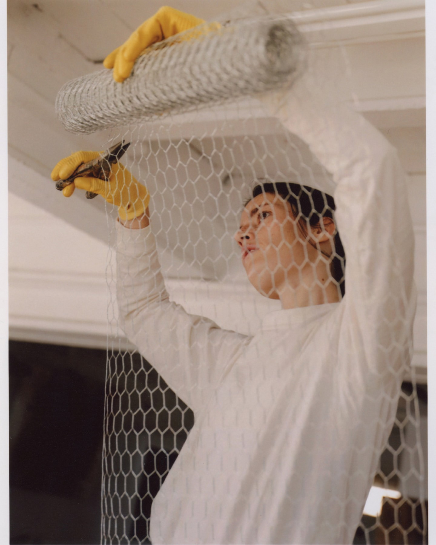 Adult working with chicken wire, using pliers, wearing yellow gloves in an indoor setting with a roll of wire.