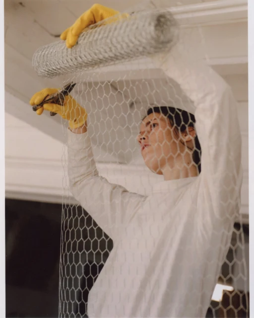 Adult working with chicken wire, using pliers, wearing yellow gloves in an indoor setting with a roll of wire.