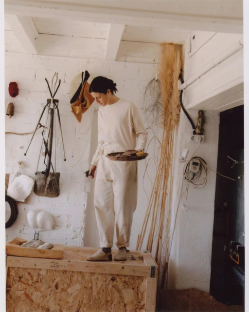 Adult female in long sleeves and pants standing on a wooden crate, holding a tray in an indoor setting with neutral and muted tones.