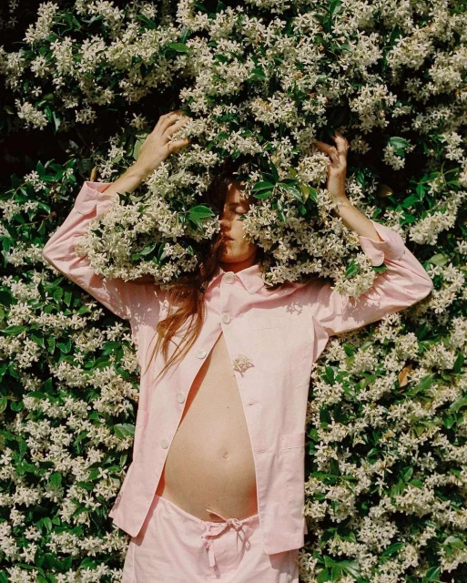 Pregnant woman in a pink pajama set surrounded by white star jasmine flowers, lying down in a garden setting.