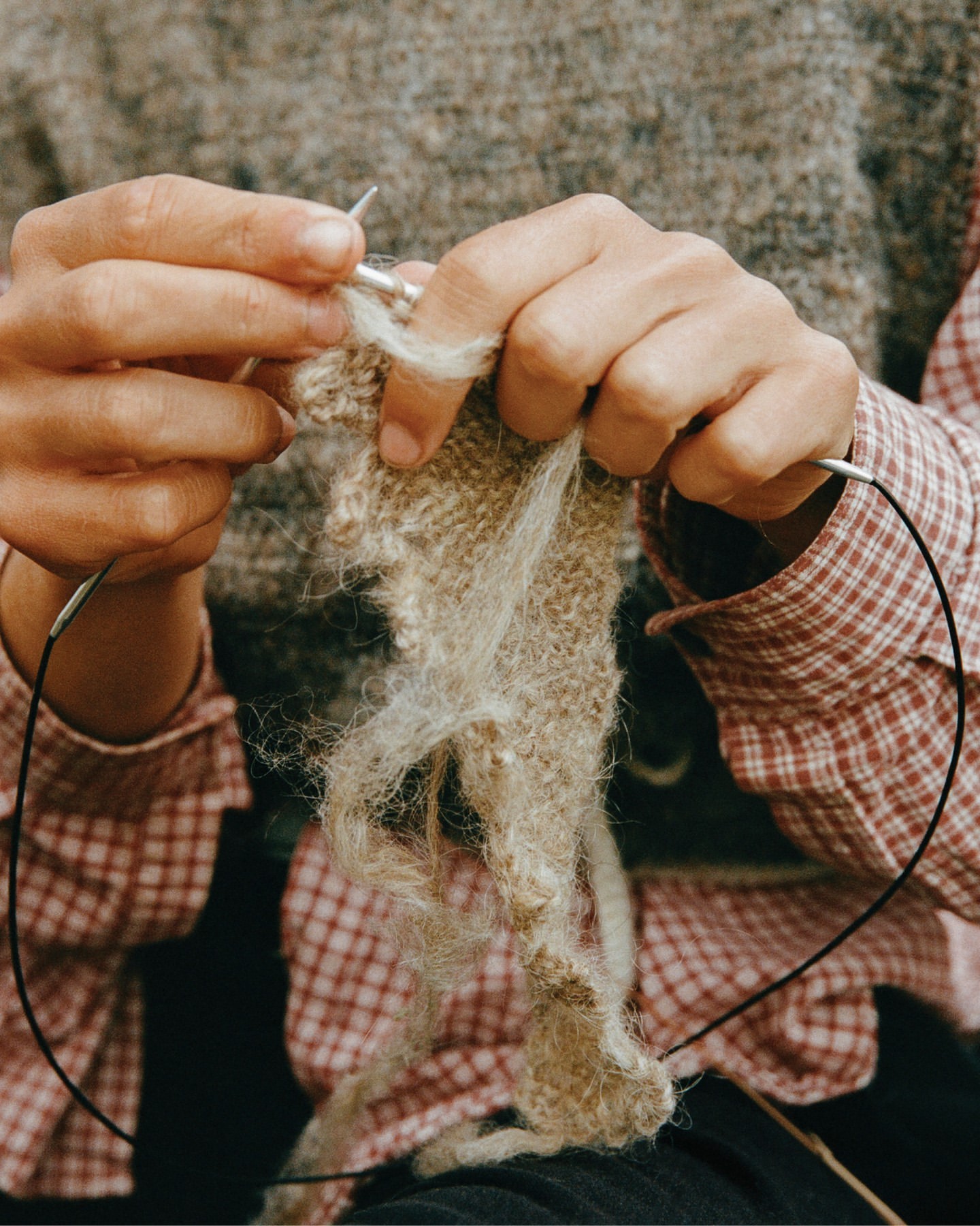 Close-up of adult hands knitting with needles and wool yarn; wearing a plaid shirt with soft, diffused light on the wool and hands.