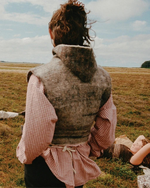 Woman in vest stands in a field, viewed from the back. Another person lies in the grass with a blue sky in the background.